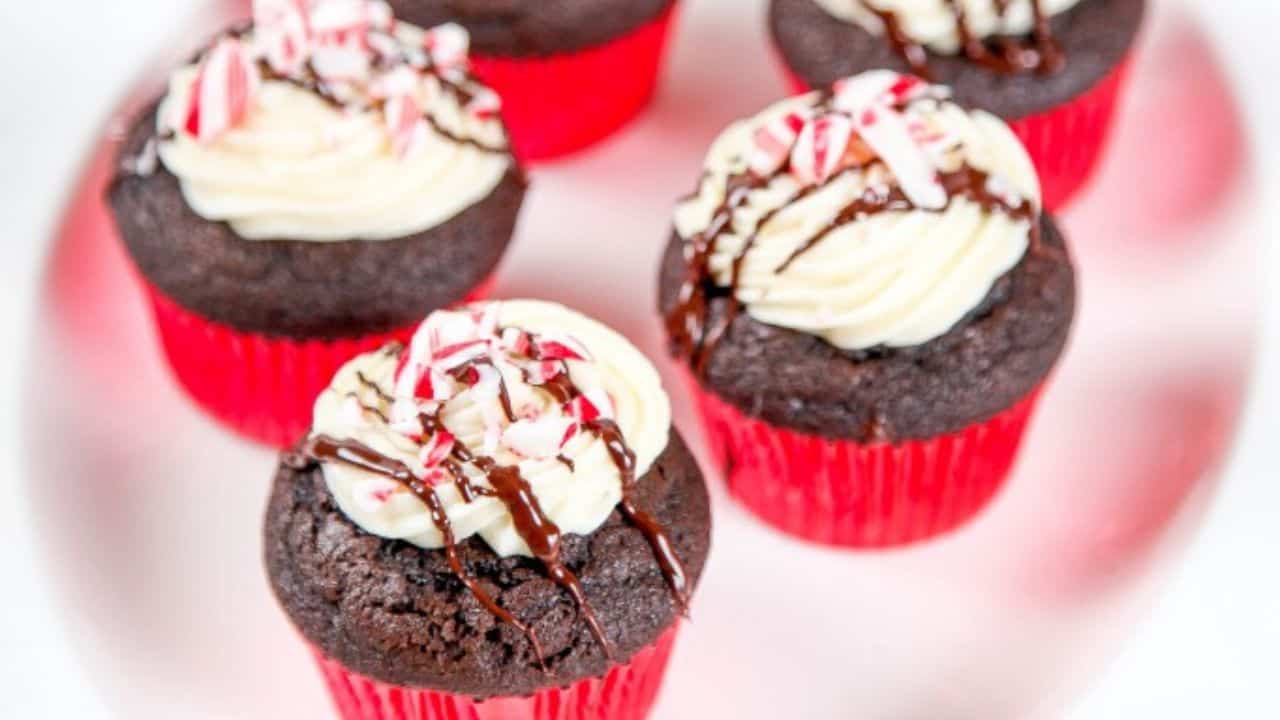 Five chocolate cupcakes with white frosting, red sprinkles, and chocolate drizzle are arranged on a white surface. Each cupcake is in a red wrapper.