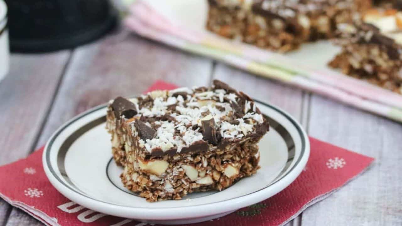 A square chocolate oat bar with shredded coconut on top sits on a small plate, placed on a red napkin with another bar visible in the background.