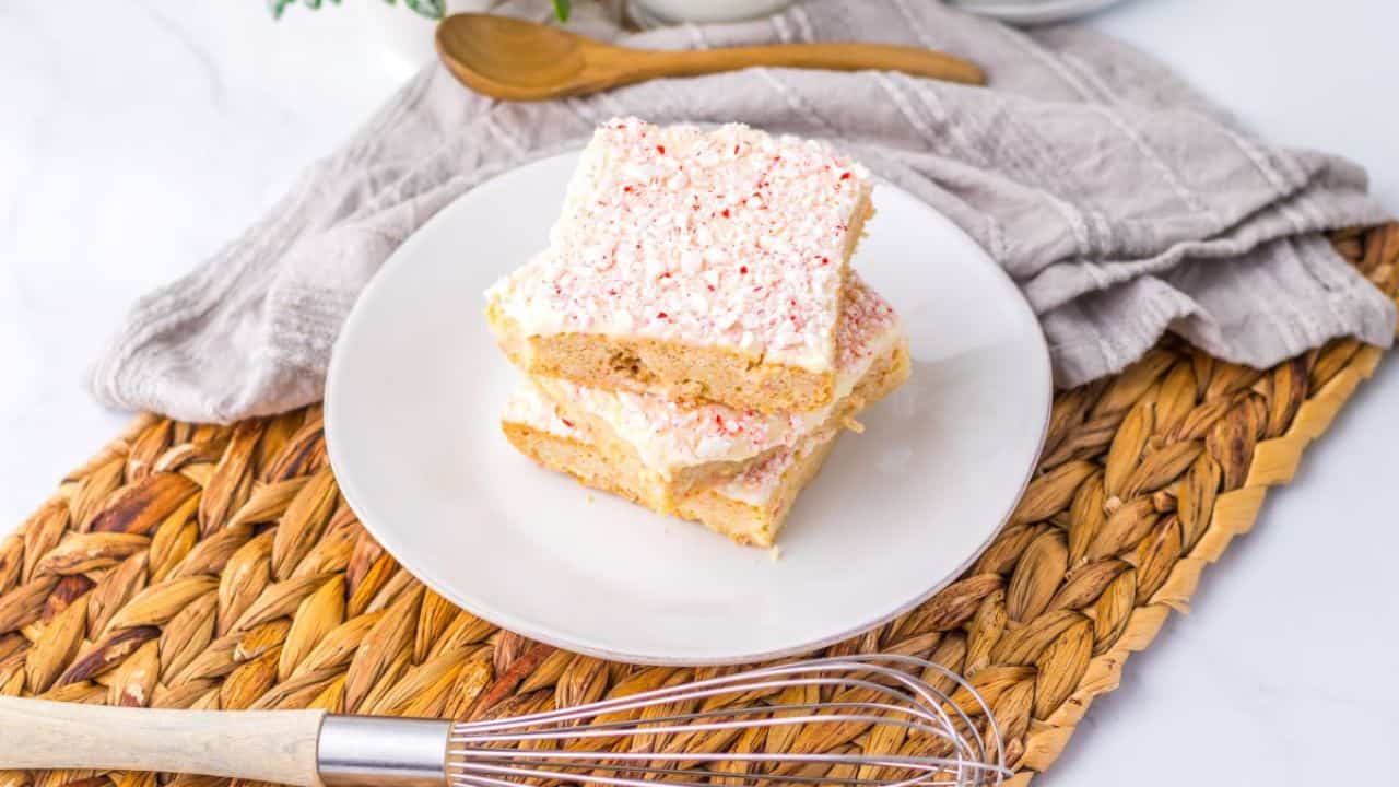 Two frosted dessert bars stacked on a white plate, placed on a woven mat with a whisk and spoon nearby.