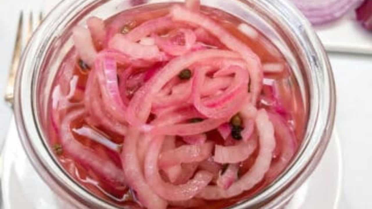 A jar of pickled red onions on a kitchen counter, with raw red onion slices and a fork nearby.