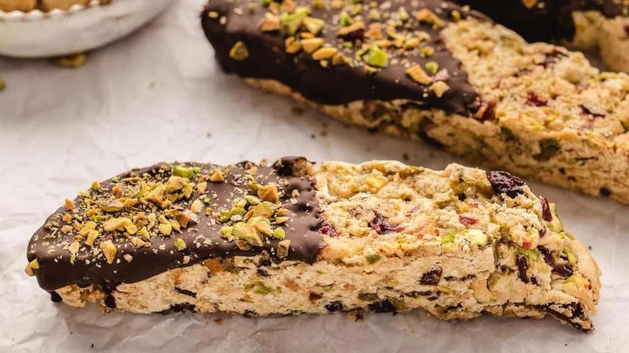 Biscotti topped with a chocolate coating and sprinkled with chopped pistachios, featuring visible nuts and dried fruit within the cookie.