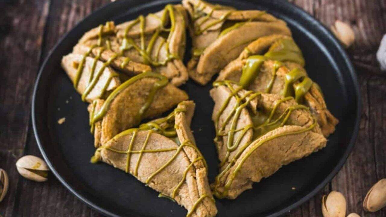 A black plate holds several triangular pistachio cookies drizzled with green icing, arranged in a circular pattern on a wooden surface.