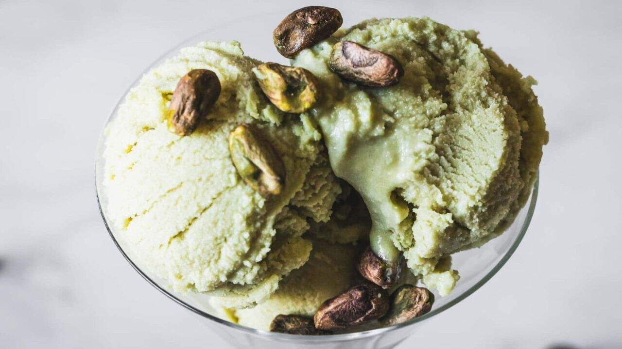 Three scoops of pistachio ice cream served in a glass dish, topped with several whole pistachio nuts, against a blurred light background.