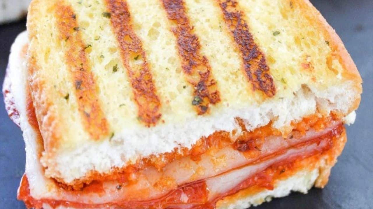 A close-up of a grilled sandwich with toasted bread, melted cheese, pepperoni, and tomato sauce, cut in half on a dark surface.