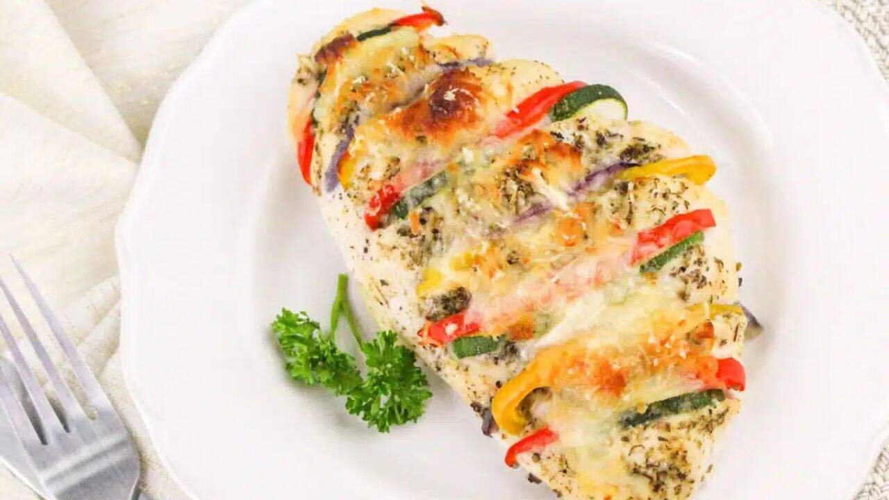 A baked chicken breast stuffed with slices of zucchini, red bell pepper, and cheese, garnished with herbs, on a white plate with a sprig of parsley.