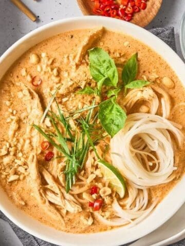 A bowl of creamy noodle soup topped with shredded chicken, fresh herbs, chopped peanuts, and sliced red chili, with lime slices and a drink nearby.