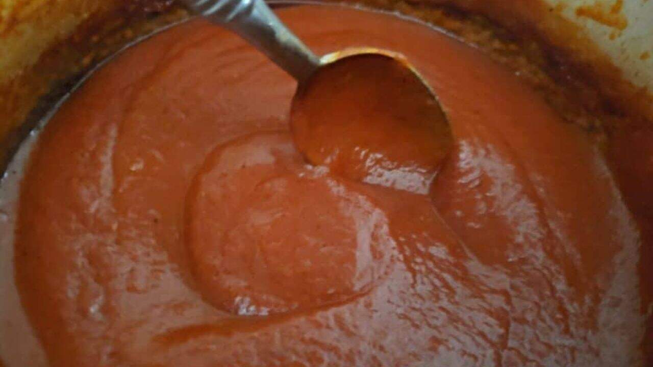 A wooden spoon has red enchilada sauce dripping off it into a blue pot filled with the remainder of the homemade enchilada sauce.
