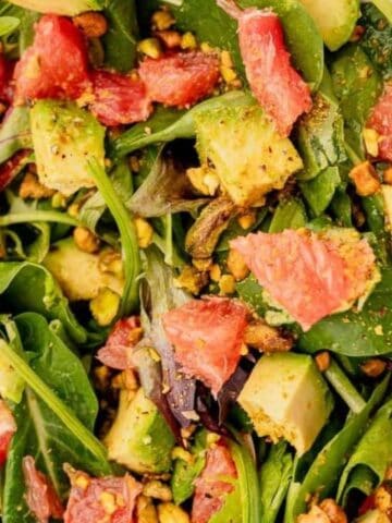 A fresh salad with spinach, avocado chunks, grapefruit pieces, and pistachios, served in a bowl with wooden serving utensils.