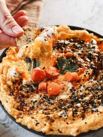 A hand dips a seeded cracker into a bowl of whipped dip topped with carrots, seeds, sauce, and parsley on a marble surface.