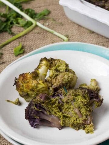 A plate with roasted broccoli and cauliflower, a fork and knife, a small dish of oil, and a baking dish with more vegetables in the background.