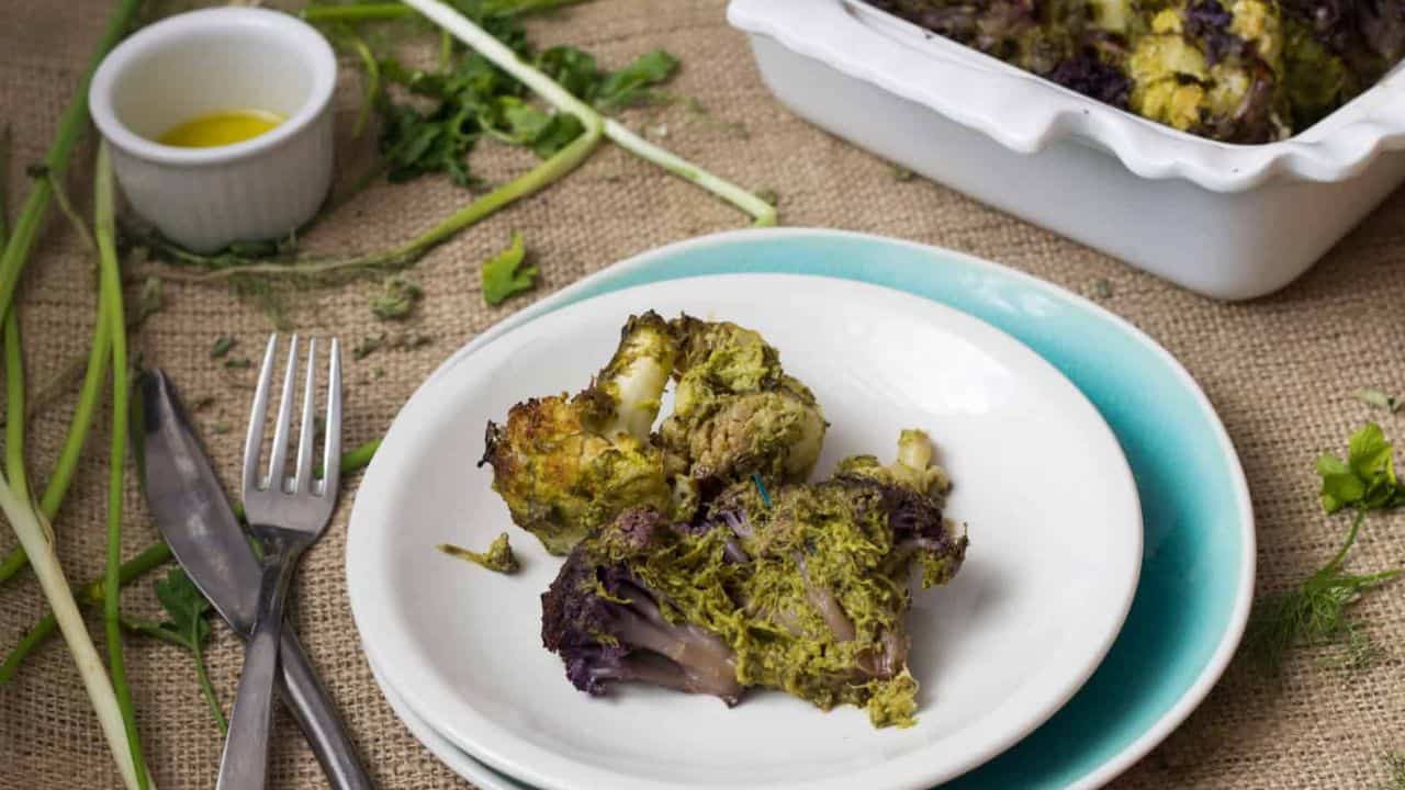 A baked dish with roasted cauliflower florets covered in a green herb sauce is displayed in a white ceramic baking dish. A sprig of fresh parsley garnishes the top. A small bowl of olive oil and a few scallions are placed beside the dish.