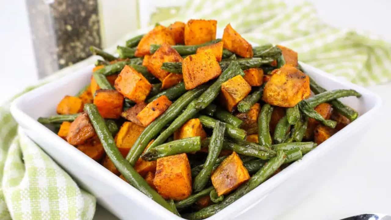A square white bowl filled with roasted sweet potatoes and green beans, placed on a green checkered cloth, with a spoon and pepper mill visible in the background.