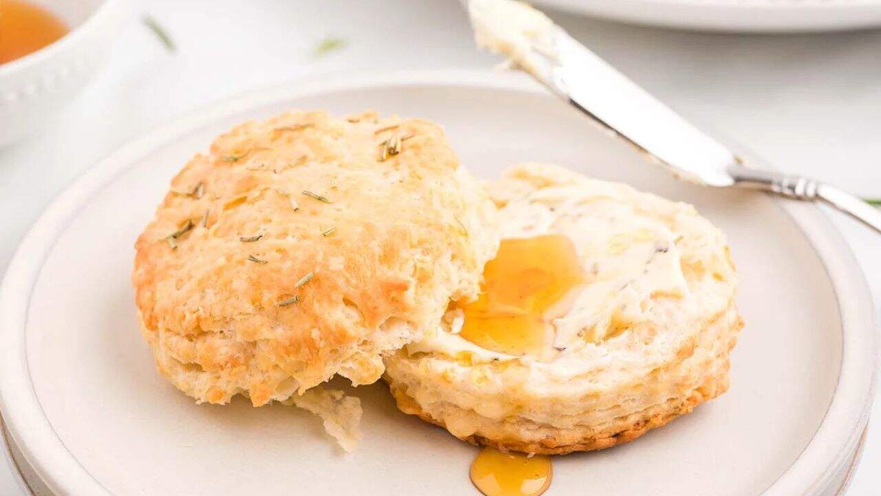 A split biscuit with butter and honey on a white plate, next to a butter knife.