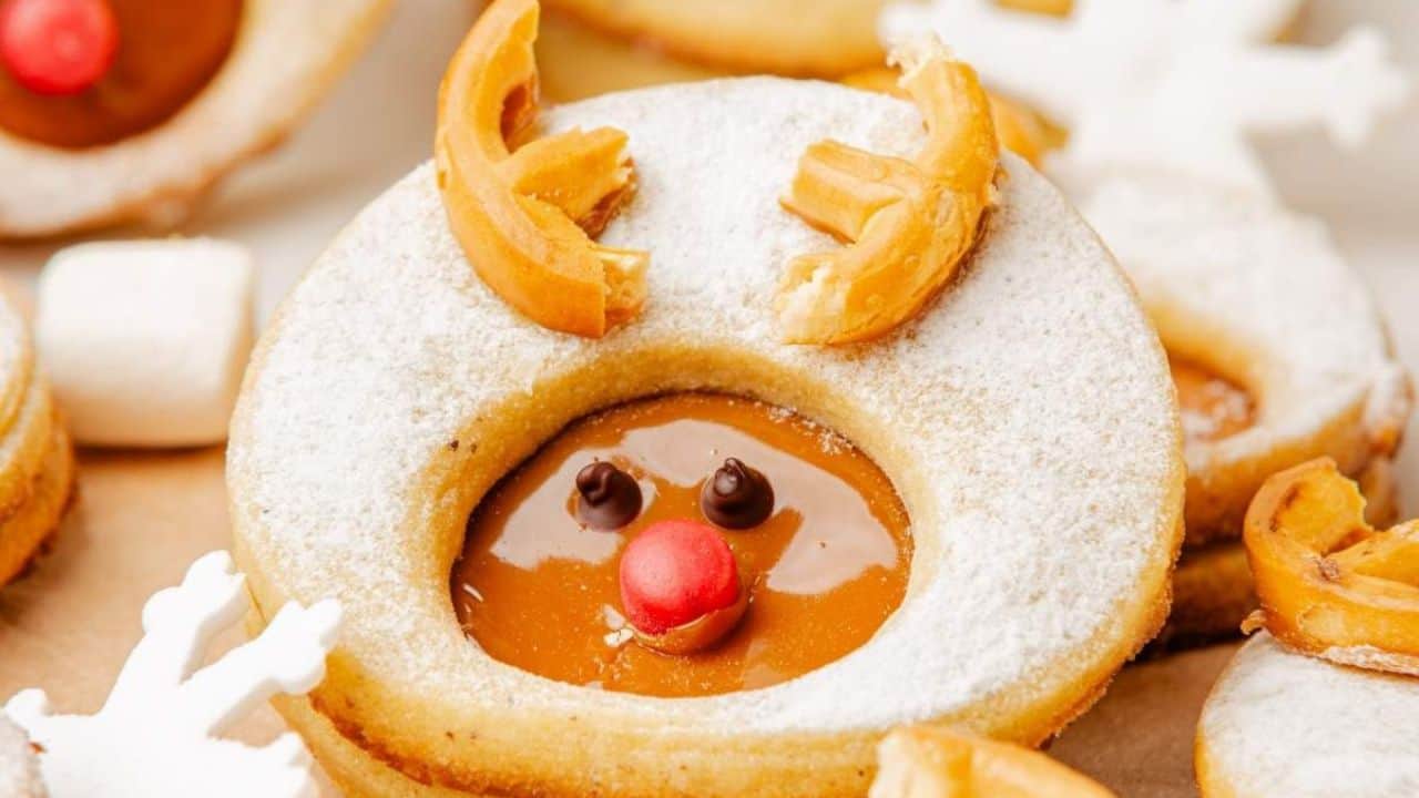 A round cookie dusted with powdered sugar, filled with caramel, decorated to look like a reindeer with pretzel antlers, chocolate eyes, and a red candy nose.