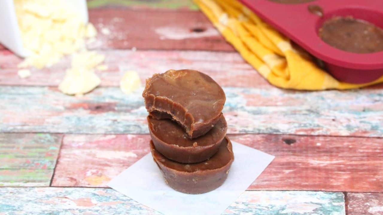Three chocolate fat bombs are stacked on parchment paper, with the top one partially bitten. A yellow cloth and silicone mold are visible in the background.