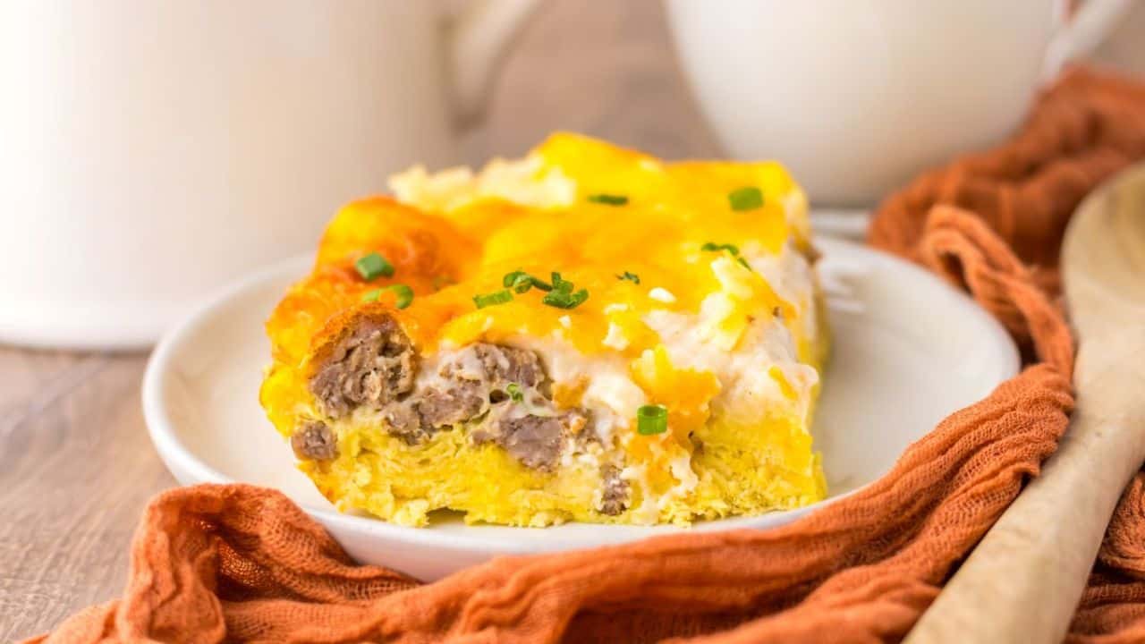 A slice of breakfast casserole with sausage, eggs, and cheese is served on a white plate beside an orange cloth and a wooden spoon.