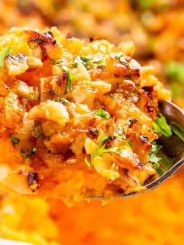 A spoonful of mashed sweet potatoes topped with a crumbly, nutty mixture, held above a baking dish containing the same casserole.