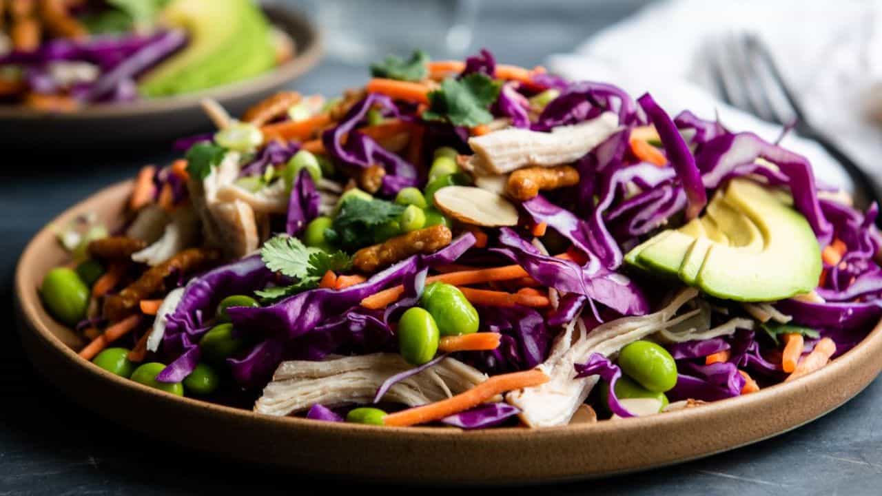 A plate of salad featuring shredded purple cabbage, carrots, avocado slices, edamame, chicken, cilantro, and nuts.