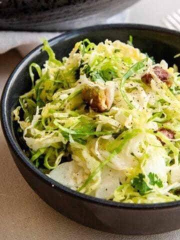 A black bowl filled with a shredded cabbage salad topped with nuts and herbs, placed on a light surface beside a napkin and utensils.