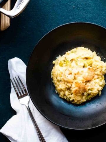 A black plate with a serving of creamy risotto on a dark surface, accompanied by a fork, white napkin, and a glass of white wine.
