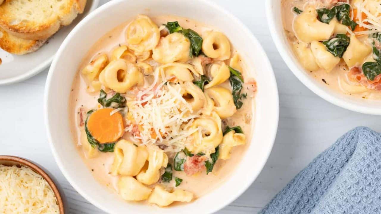 A bowl of creamy tortellini soup with spinach, carrots, and shredded cheese on top, served next to bread slices and a dish of grated cheese.