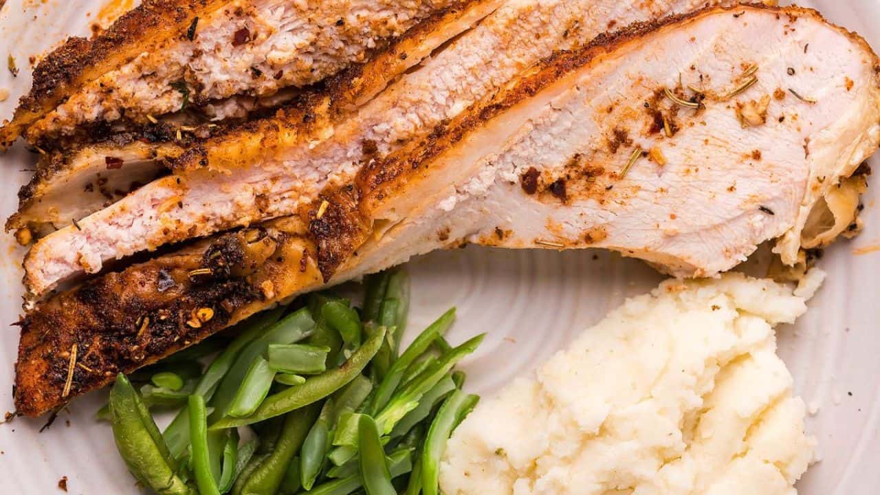 Sliced seasoned roast turkey breast served with mashed potatoes and green beans on a white plate.