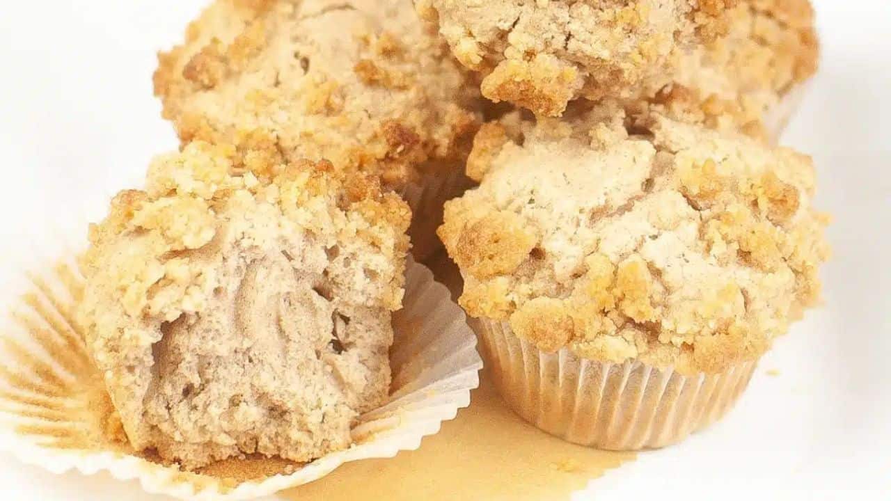 A plate of five cinnamon muffins with crumbly tops; one muffin partially unwrapped and cut in half.