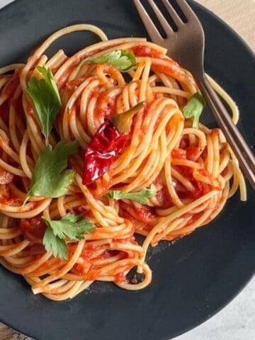 A black plate of spaghetti with tomato sauce, garnished with fresh herbs and a red chili, sits on a wooden board with a fork on the side.