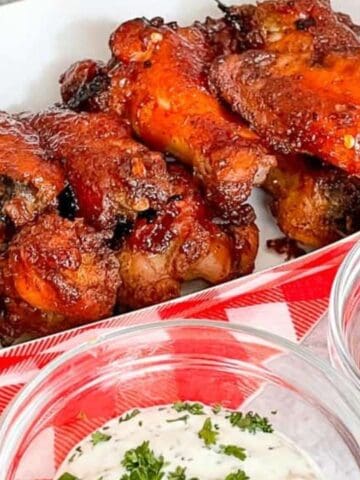 A serving tray with several glazed chicken wings, accompanied by two small bowls of dipping sauces, one white with herbs and one red, on a red and white checkered cloth.