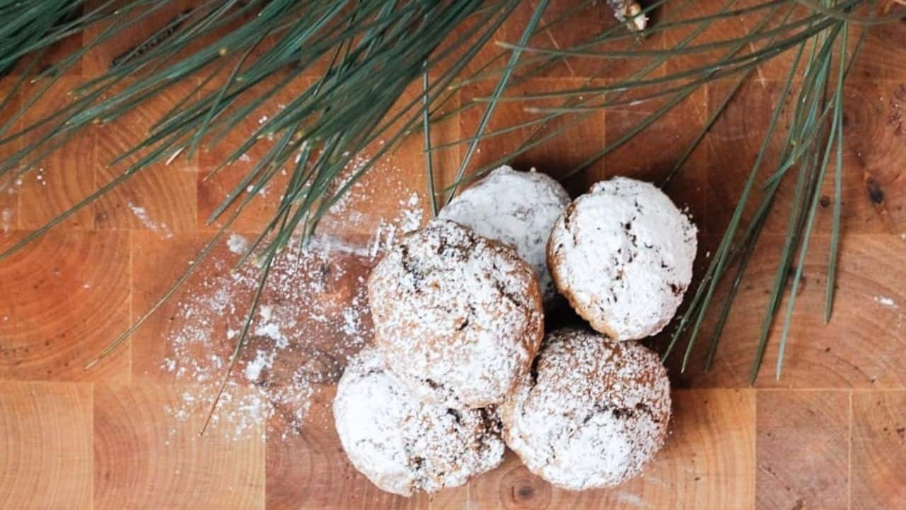 Four powdered sugar-covered cookies on a wooden surface with pine needles nearby.