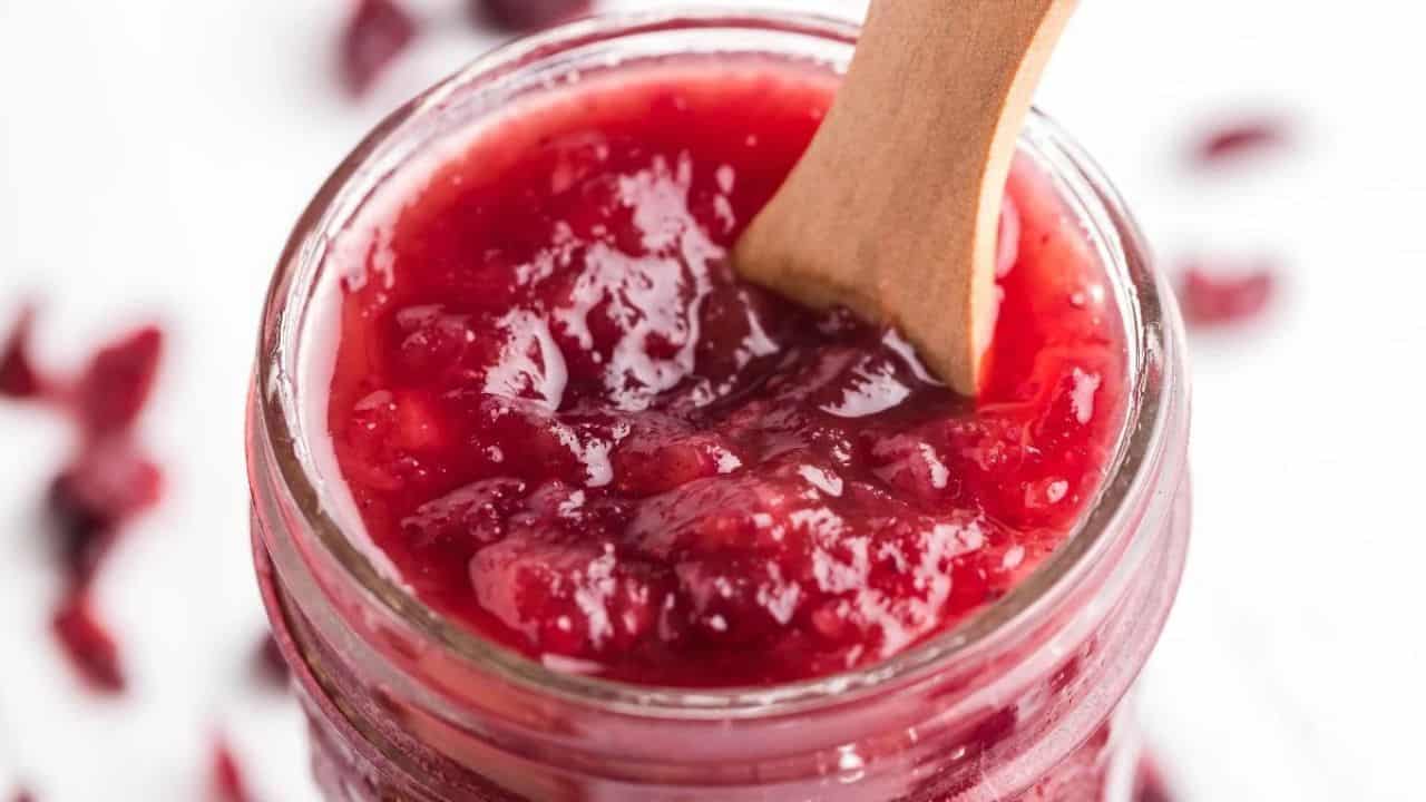 A close-up of a glass jar filled with red cranberry sauce, with a wooden spoon resting inside.