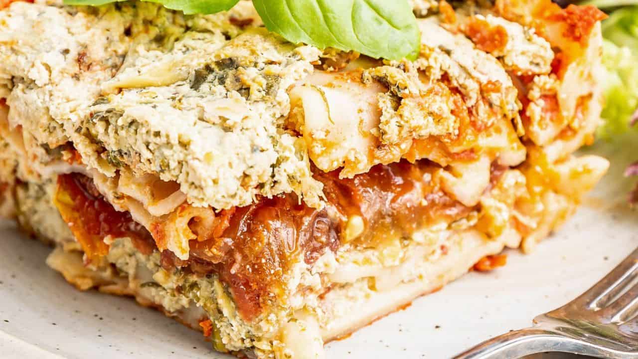 A close-up of a slice of lasagna with layers of pasta, tomato sauce, cheese, and herbs on a white plate next to a fork.