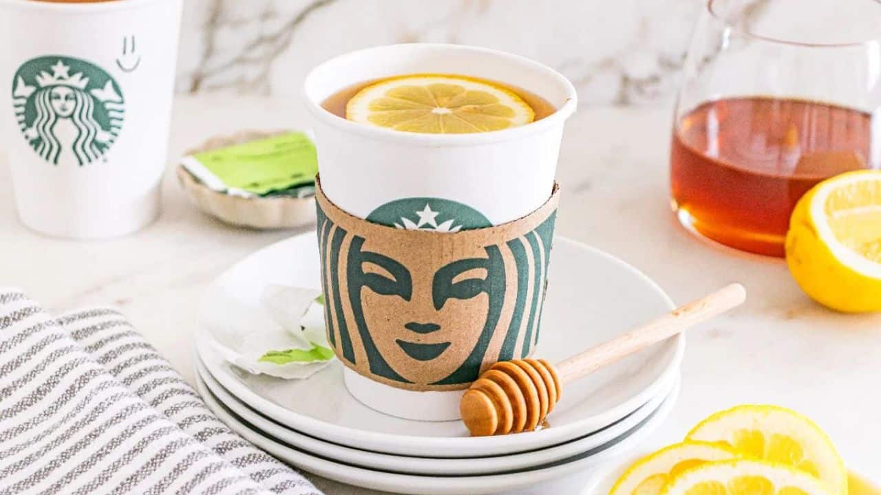 A Starbucks cup with a lemon slice in tea sits on a plate with a honey dipper, tea bag, sliced lemon, and a carafe of tea in the background.
