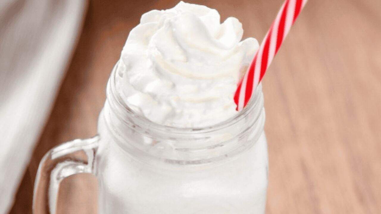 A glass jar filled with a white milkshake topped with whipped cream, served with a red and white striped straw on a wooden surface.