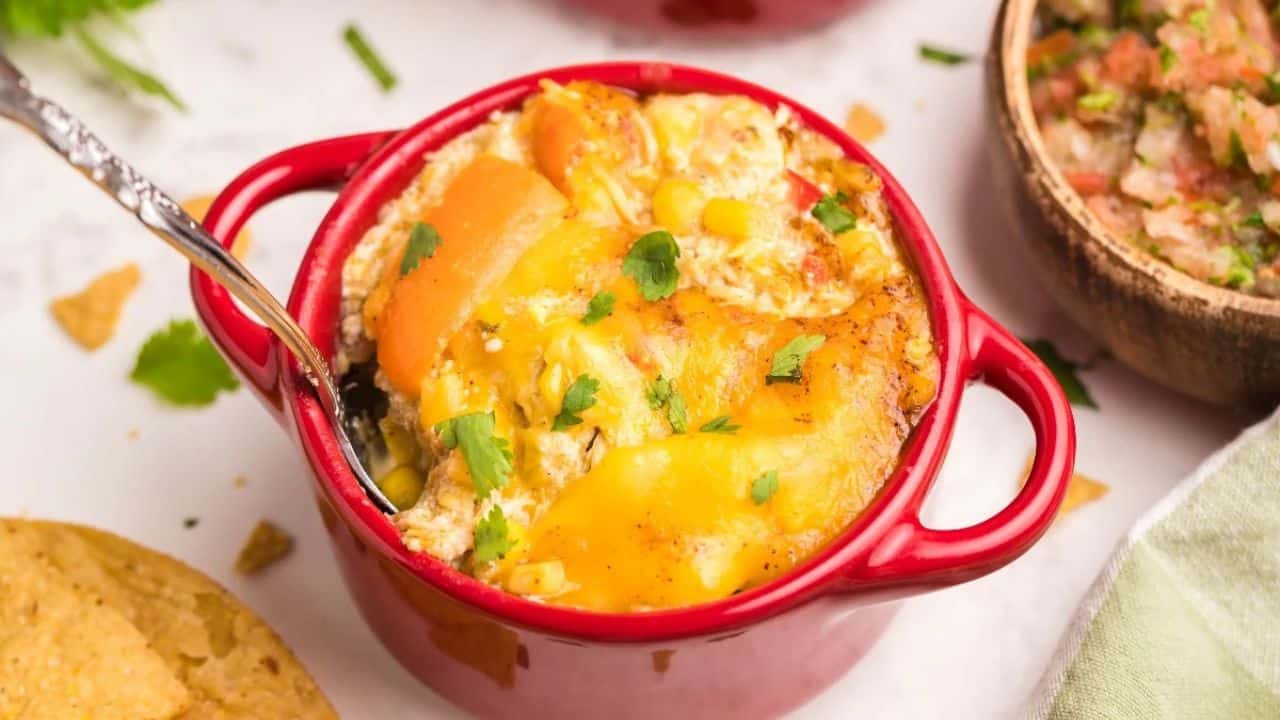 A red ramekin filled with a baked cheesy casserole topped with herbs, with a metal spoon inside, and a bowl of salsa and tortilla chips nearby.