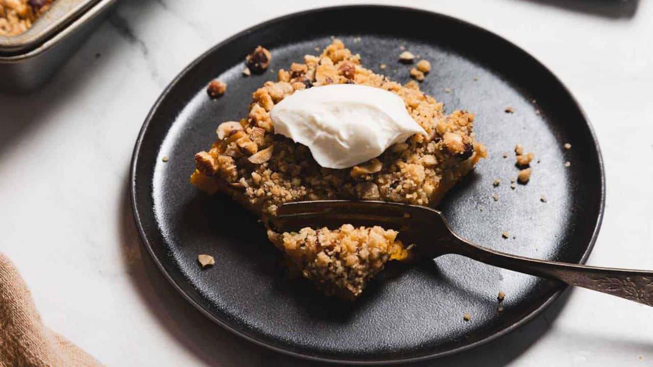A slice of crumbly dessert topped with a dollop of white cream is presented on a dark plate. The dessert is garnished with chopped nuts, and a fork rests in front of it, partially obscuring the dessert.