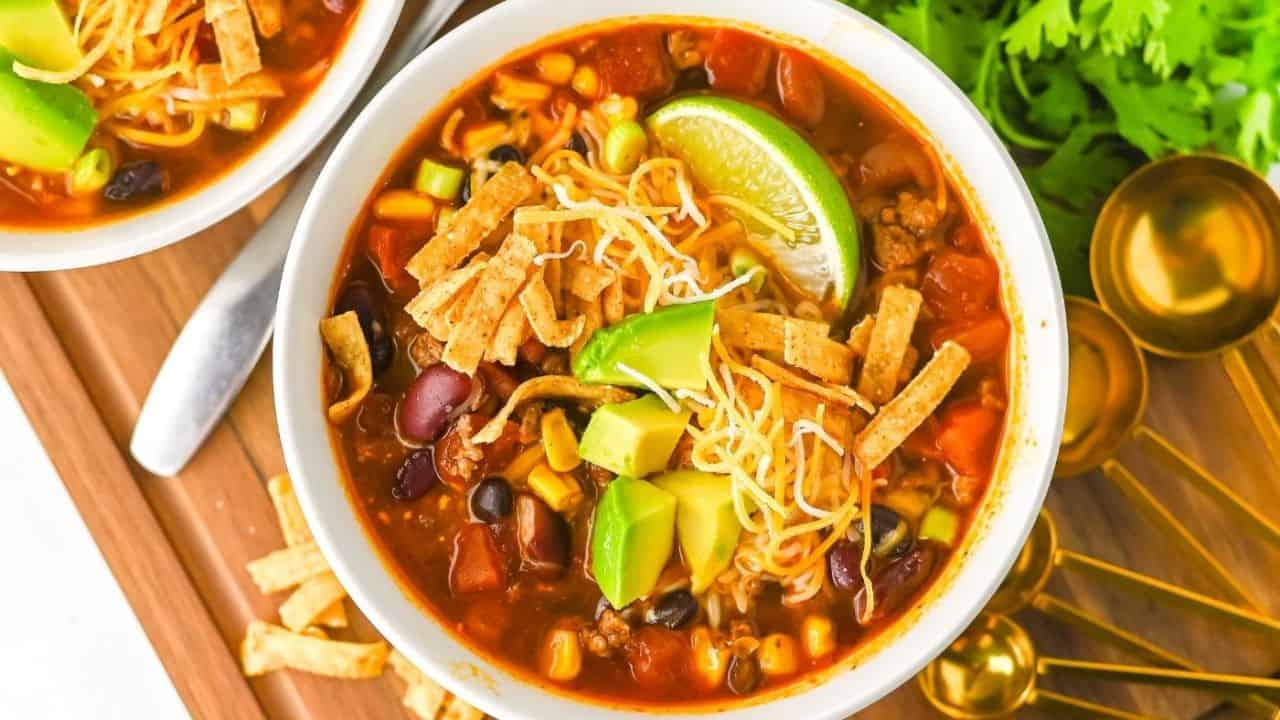 A bowl of taco soup topped with shredded cheese, tortilla strips, avocado cubes, and a lime wedge. Visible ingredients include beans, corn, tomatoes, and ground meat. A bunch of cilantro and gold measuring spoons are nearby.