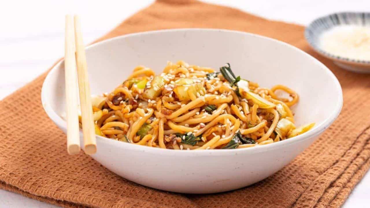 A bowl of stir-fried noodles with vegetables, topped with sesame seeds, placed on a brown cloth napkin. Chopsticks rest on the bowl's edge, and a small dish is visible in the background.