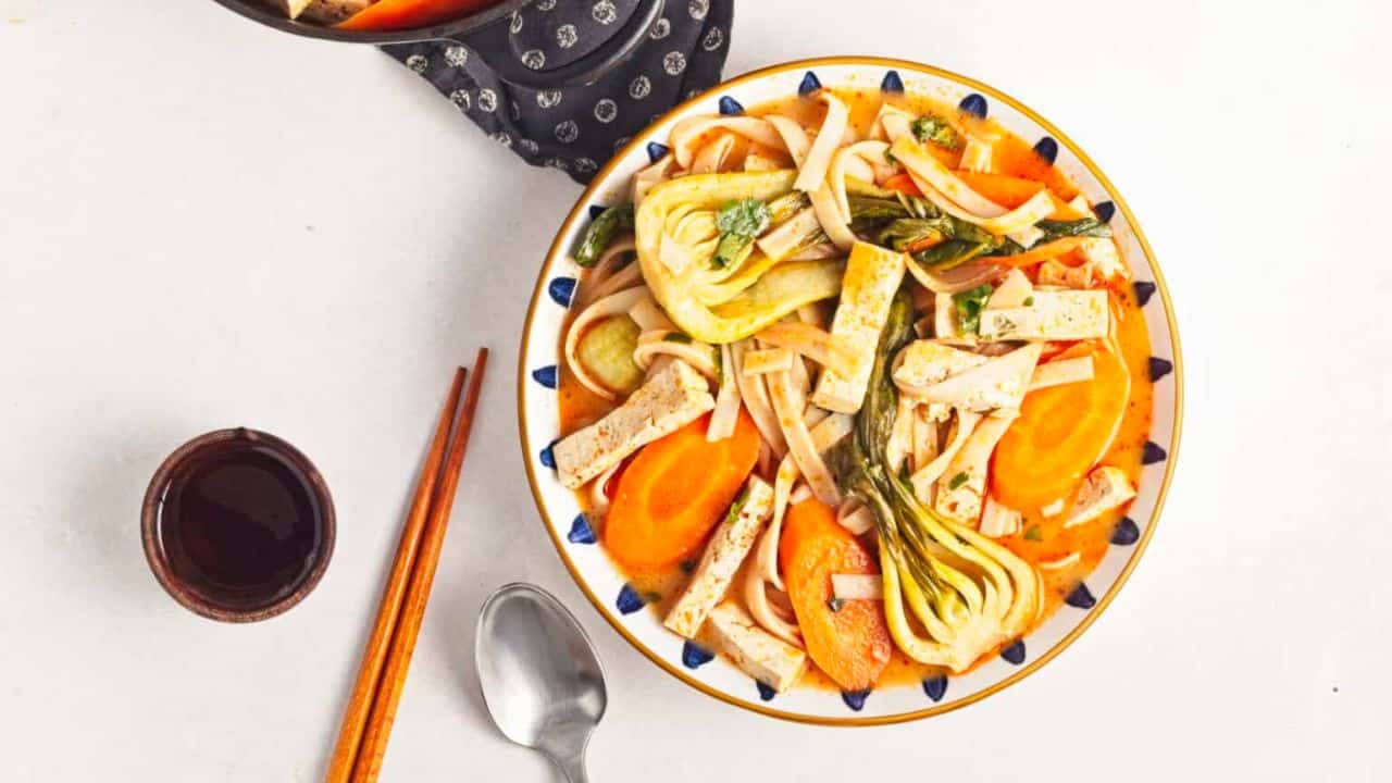 A bowl of noodle soup with tofu slices, baby bok choy, carrot rounds, and fresh herbs in a light, orange-colored broth.