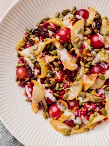 A white plate contains a salad with red grapes, sliced pears, pomegranate seeds, pepitas, and a creamy dressing, with a spoon resting on the plate.