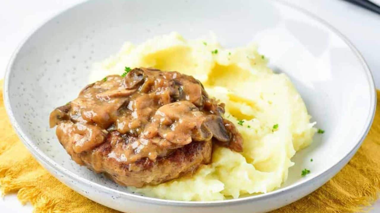 A plate with mashed potatoes topped with a turkey Salisbury steak and mushroom gravy, placed on a yellow cloth napkin. A fork is in the background.