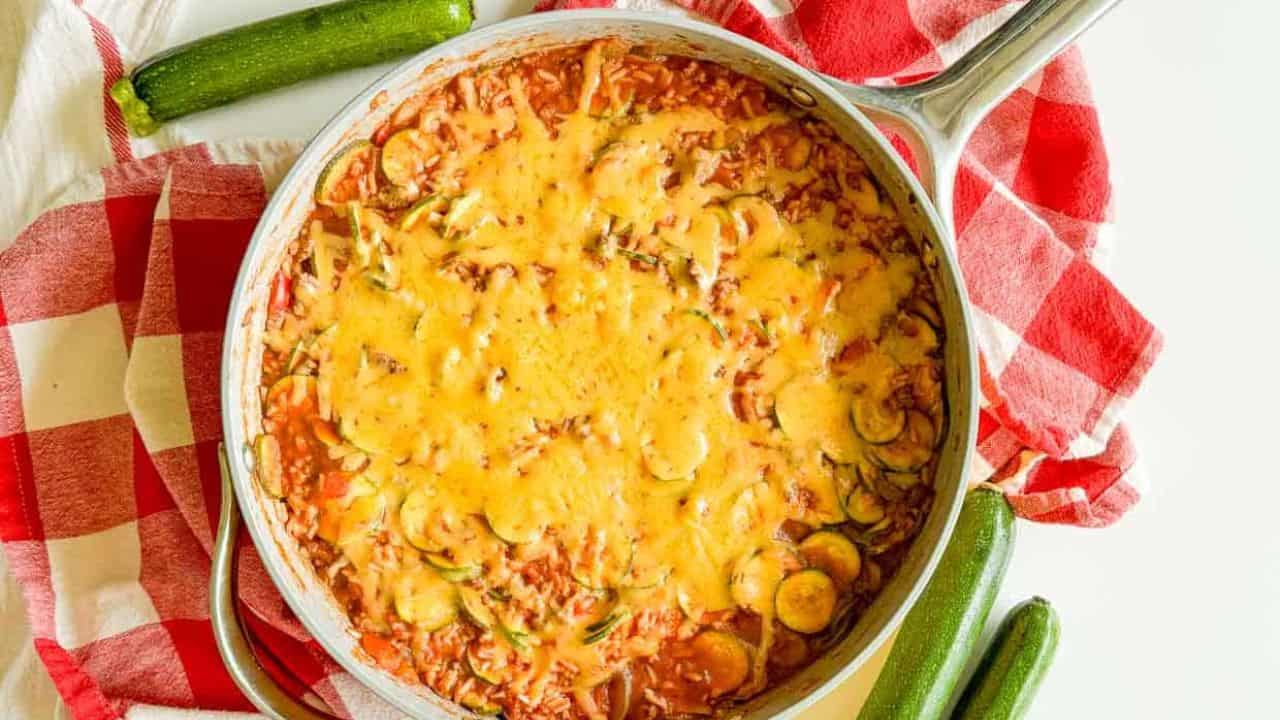 A pan of cheese-topped unstuffed zucchini casserole on a red and white checkered cloth with three whole zucchinis placed around it.