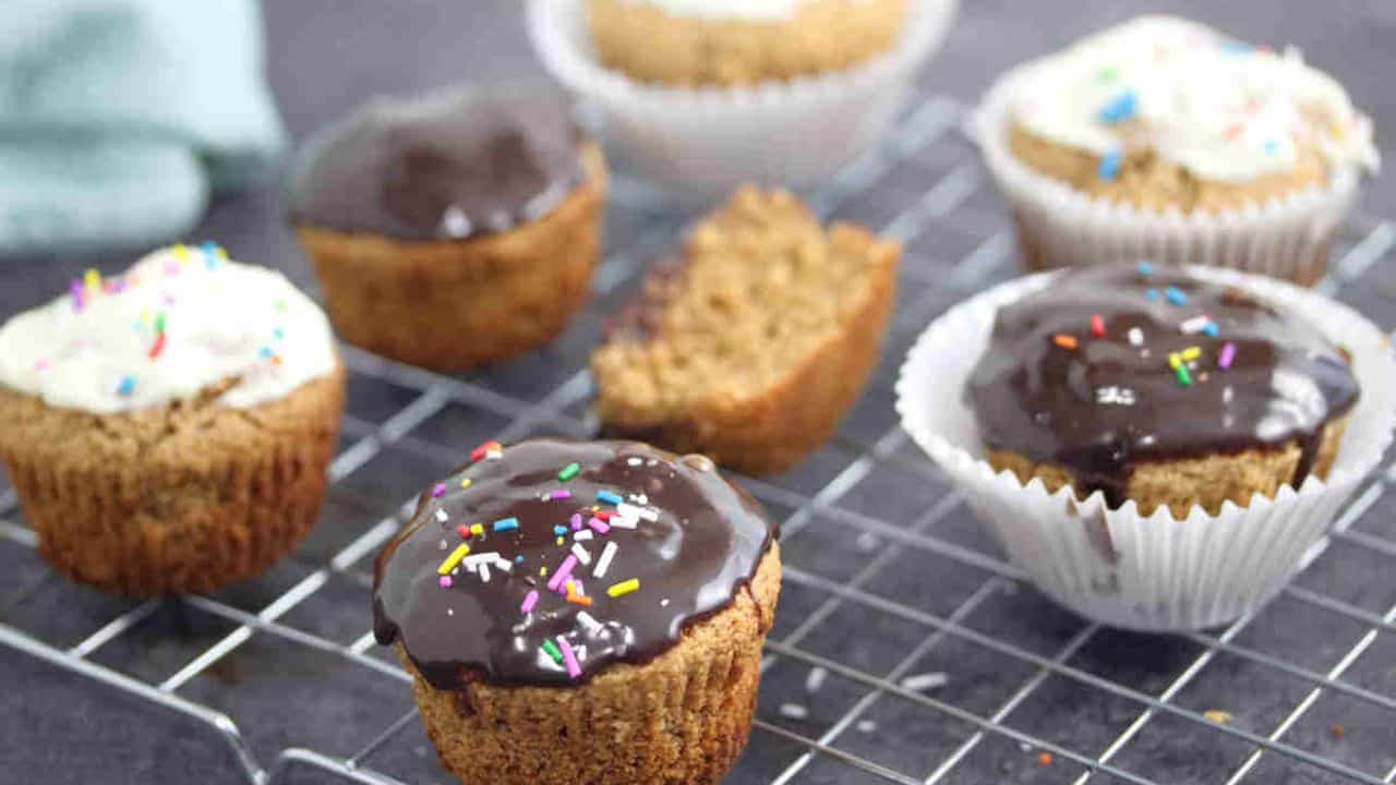 Cupcakes with chocolate and vanilla frosting, some topped with sprinkles, are displayed on a cooling rack, with one cupcake cut in half.