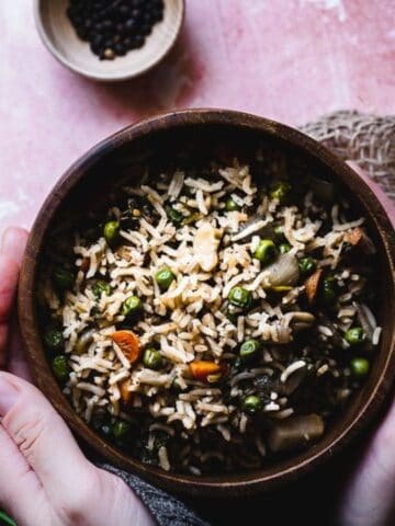 A person holds a bowl of rice mixed with vegetables; nearby are a spoon, fresh cilantro, cashews, and a bowl of black peppercorns on a pink textured surface.