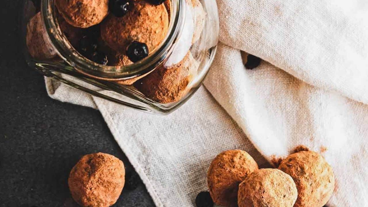 Chocolate truffles coated with cocoa powder are placed on a gray surface. Some of the truffles and small dried berries are in the background, slightly out of focus. A light fabric is draped behind them.