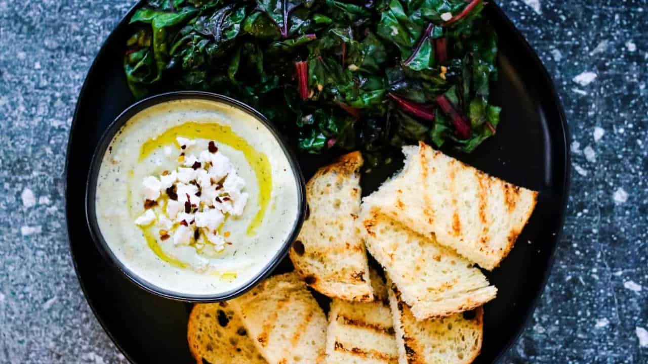 Overhead shot of a black platter with a bowl of whipped feta spread, sauteed chard, and grilled sourdough bread.