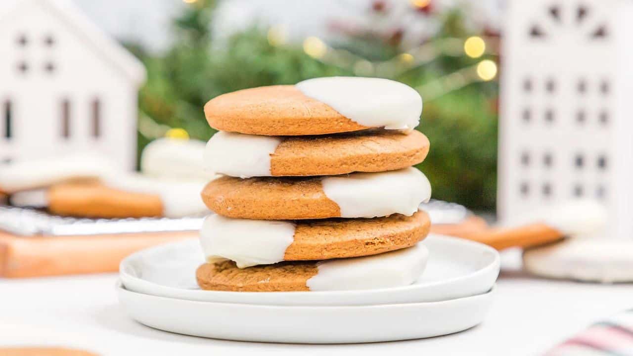 A stack of four round cookies half-dipped in white icing sits on a white plate, with festive decorations blurred in the background.