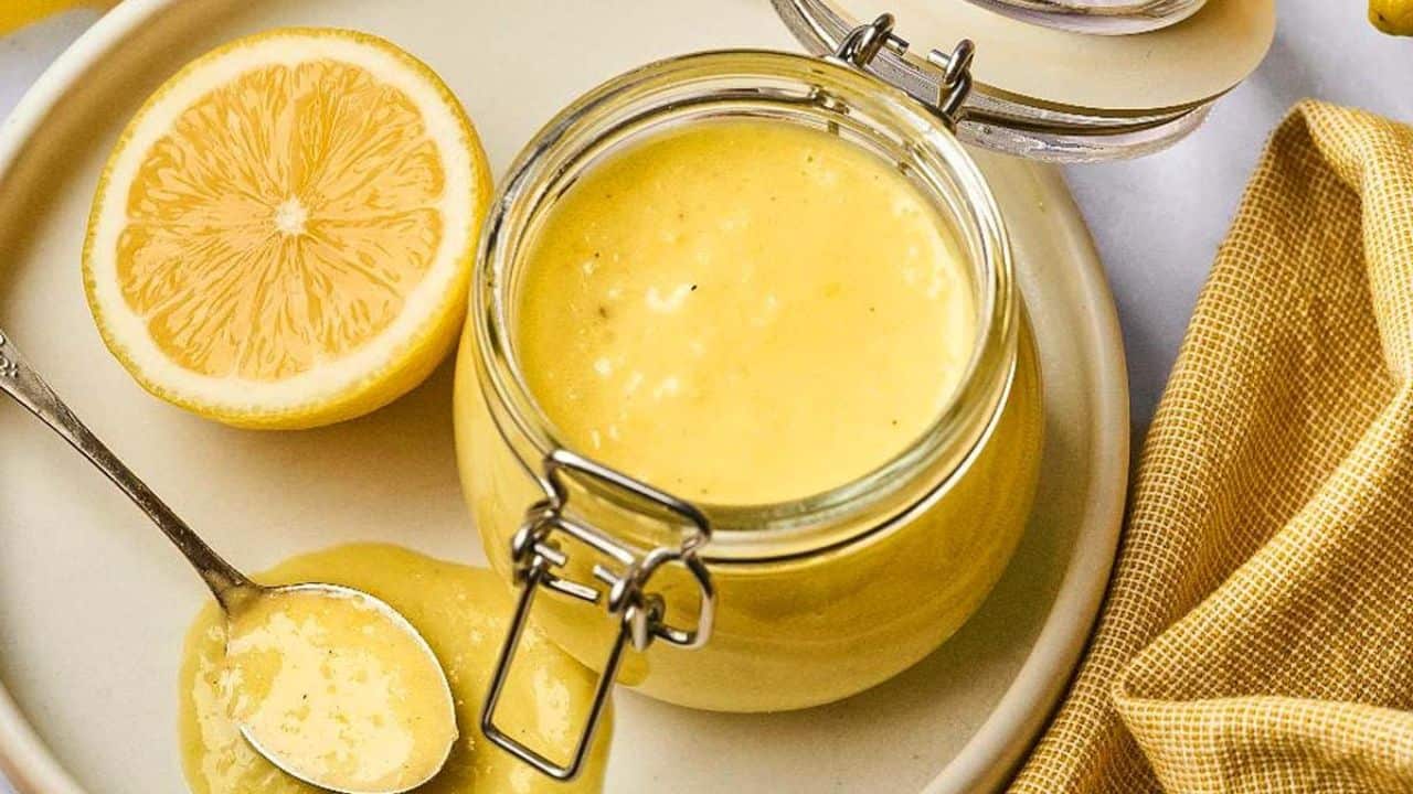 An open jar of lemon curd sits on a plate with a halved lemon, a spoonful of curd, and a yellow napkin beside it.