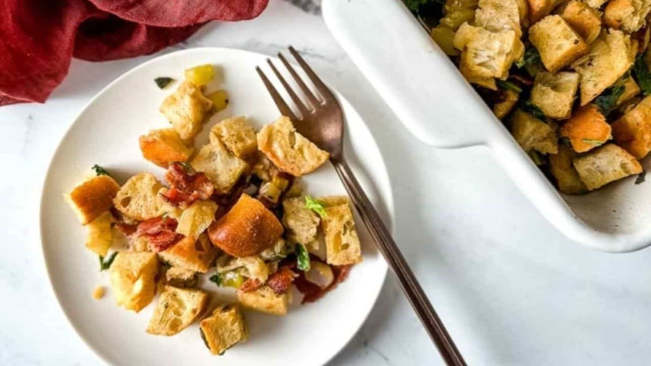 A plate with crouton stuffing, pieces of bread, and herbs beside a fork; a serving dish with more stuffing is nearby on a marble surface.