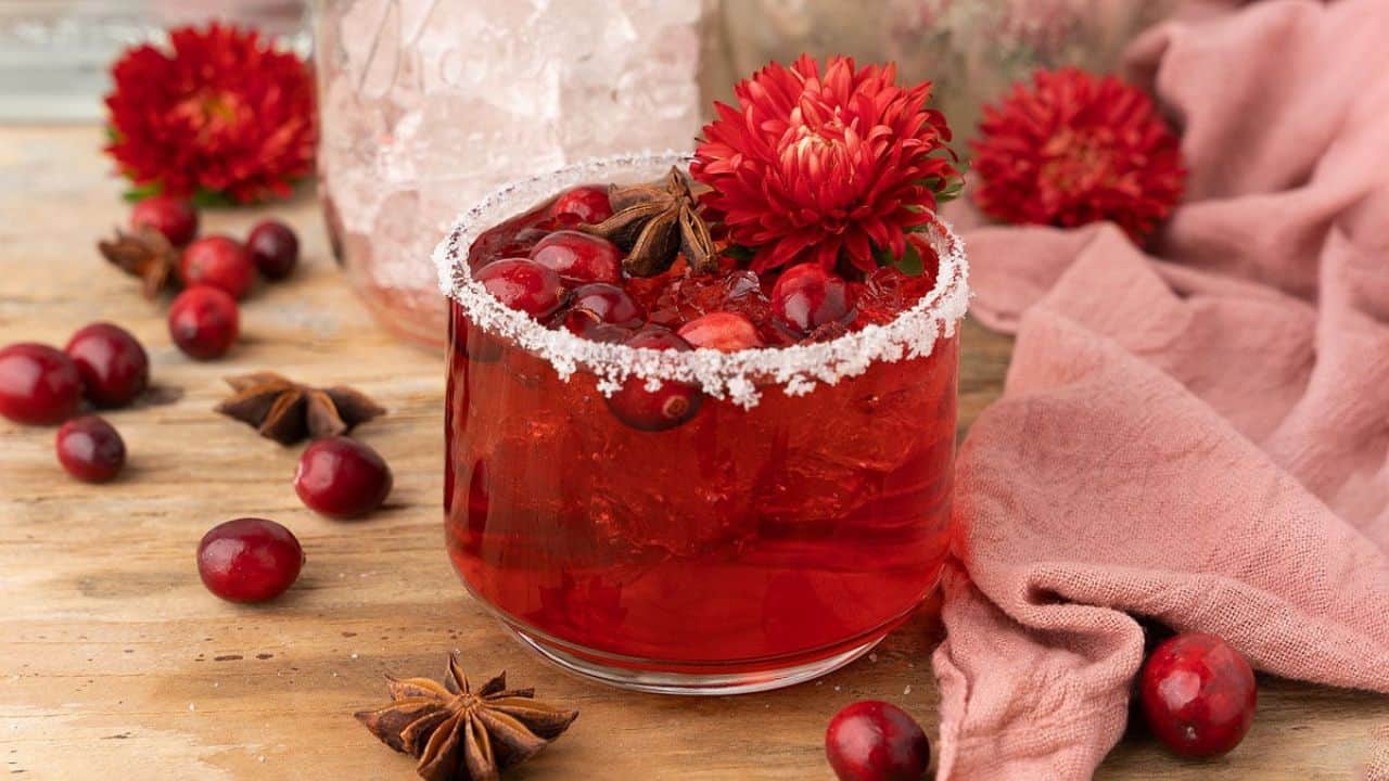 A glass of red cranberry drink with ice, sugared rim, star anise, cranberries, and a red flower garnish sits on a wooden surface with scattered cranberries and fabric nearby.